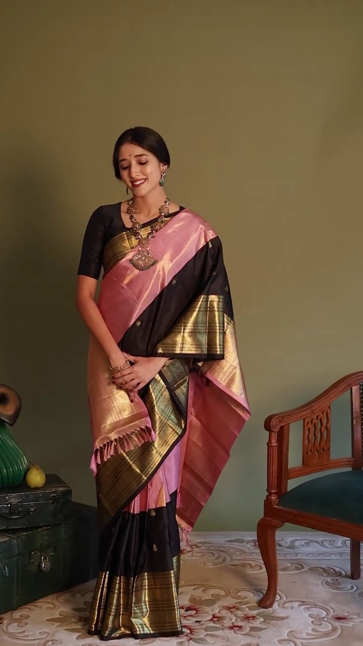 Woman in a traditional saree with a gold and pink pattern, standing in a room with a wooden chair and decorative items.